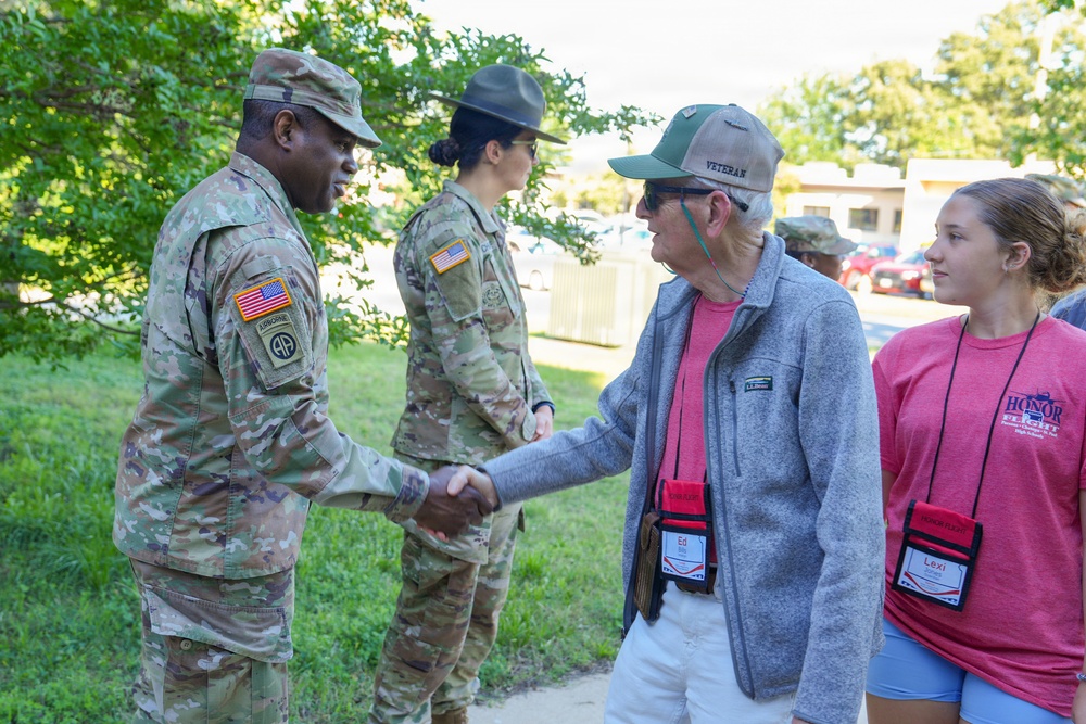 A U.S. Army soldier wearing an 82nd Airborne Division patch shakes hands with a civilian veteran wearing a 'VETERAN' cap and a name badge reading 'Ed'. A young woman named 'Lexi' wearing an 'Honor Flight' t-shirt stands nearby, along with a second soldier in the background wearing a drill sergeant hat. The setting appears to be an outdoor military installation or event.