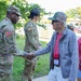 A U.S. Army soldier wearing an 82nd Airborne Division patch shakes hands with a civilian veteran wearing a 'VETERAN' cap and a name badge reading 'Ed'. A young woman named 'Lexi' wearing an 'Honor Flight' t-shirt stands nearby, along with a second soldier in the background wearing a drill sergeant hat. The setting appears to be an outdoor military installation or event.