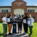 Six women are posing for a group photo in front of a brick building labeled 'Unaccompanied Housing 562,' likely on a military installation. Several of the women are holding framed award certificates or plaques, suggesting this is an award recognition ceremony. The setting appears to be outdoors on a sunny day with well-maintained grass surrounding the building.