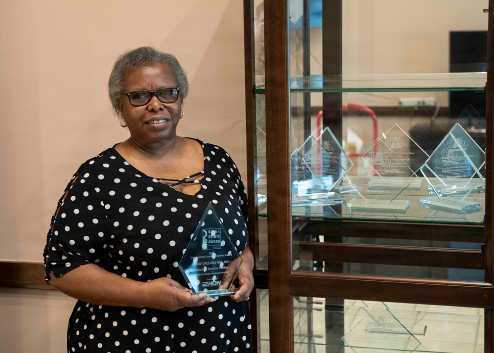 A woman wearing a black and white polka dot dress is holding a glass crystal award trophy. She is standing next to a glass display cabinet filled with multiple similar diamond-shaped glass awards. The award she holds appears to be a 'Real Estate Award' associated with 'New Orleans NAREB Service Center'.