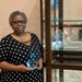 A woman wearing a black and white polka dot dress is holding a glass crystal award trophy. She is standing next to a glass display cabinet filled with multiple similar diamond-shaped glass awards. The award she holds appears to be a 'Real Estate Award' associated with 'New Orleans NAREB Service Center'.