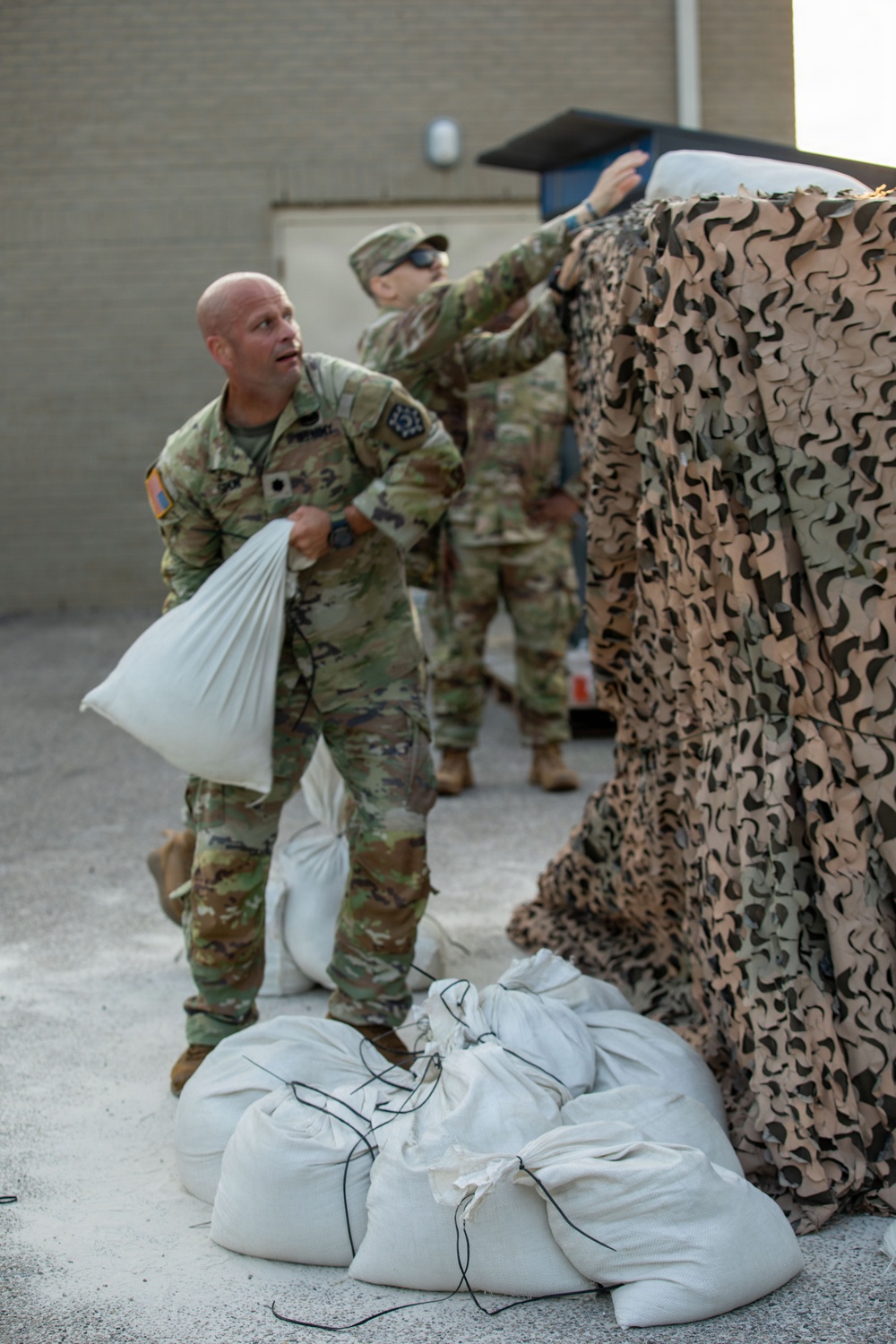 Two soldiers in US Army OCP (Operational Camouflage Pattern) uniforms are working with sandbags and a camouflage netting near a building. The foreground soldier, who is bald, is carrying a white sandbag while looking upward, and a second soldier in the background is adjusting or deploying the camouflage netting over what appears to be a vehicle or structure. Multiple tied white sandbags are visible on the ground.