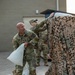 Two soldiers in US Army OCP (Operational Camouflage Pattern) uniforms are working with sandbags and a camouflage netting near a building. The foreground soldier, who is bald, is carrying a white sandbag while looking upward, and a second soldier in the background is adjusting or deploying the camouflage netting over what appears to be a vehicle or structure. Multiple tied white sandbags are visible on the ground.