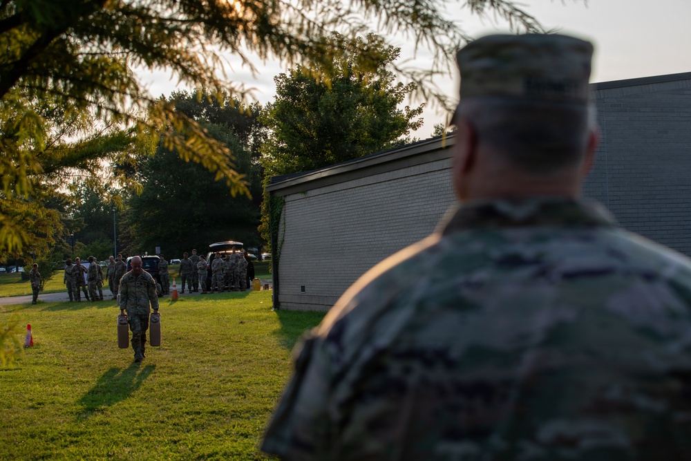 Military personnel in camouflage uniforms are gathered outdoors on a grassy area, likely during early morning or evening based on the warm, low-angle lighting. A soldier in the foreground observes the scene from behind, while another soldier in the middle ground carries what appear to be propane tanks or similar cylindrical containers. A larger group of uniformed personnel is assembled in the background near a vehicle.