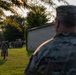 Military personnel in camouflage uniforms are gathered outdoors on a grassy area, likely during early morning or evening based on the warm, low-angle lighting. A soldier in the foreground observes the scene from behind, while another soldier in the middle ground carries what appear to be propane tanks or similar cylindrical containers. A larger group of uniformed personnel is assembled in the background near a vehicle.