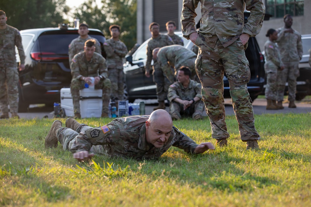 A group of military personnel in OCP (Operational Camouflage Pattern) uniforms are gathered in an outdoor area near parked vehicles. One soldier in the foreground is lying prone on the grass, appearing to be performing a low crawl exercise or drill. Other soldiers are observing, resting, or standing in the background.