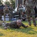 A group of military personnel in OCP (Operational Camouflage Pattern) uniforms are gathered in an outdoor area near parked vehicles. One soldier in the foreground is lying prone on the grass, appearing to be performing a low crawl exercise or drill. Other soldiers are observing, resting, or standing in the background.