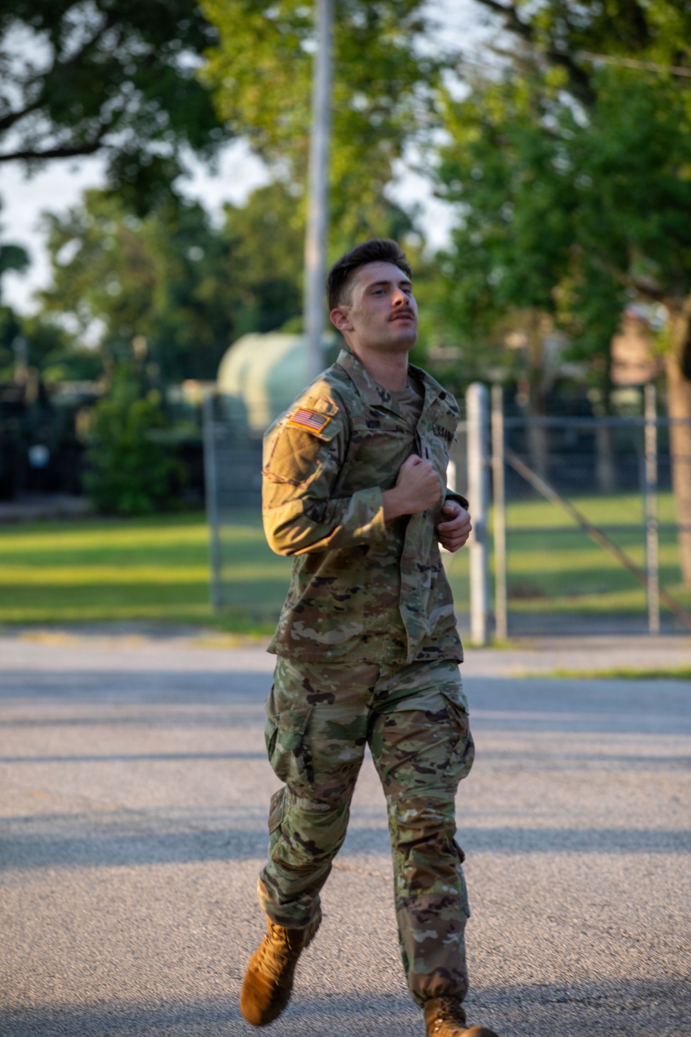 A young male U.S. Army soldier in OCP (Operational Camouflage Pattern) uniform is running or jogging on a paved road on what appears to be a military installation. He is holding his uniform jacket closed at the chest and looking upward. The setting appears to be during golden hour lighting with trees, a chain-link fence, and military equipment visible in the background.
