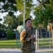 A young male U.S. Army soldier in OCP (Operational Camouflage Pattern) uniform is running or jogging on a paved road on what appears to be a military installation. He is holding his uniform jacket closed at the chest and looking upward. The setting appears to be during golden hour lighting with trees, a chain-link fence, and military equipment visible in the background.