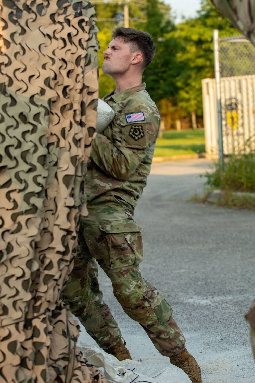 A male service member in US Army OCP (Operational Camouflage Pattern) uniform is physically handling or pulling a large camouflage netting, appearing to exert significant effort. He wears a US flag patch and a unit patch on his left sleeve. The setting appears to be a paved outdoor area near a chain-link fence with trees in the background.