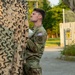 A male service member in US Army OCP (Operational Camouflage Pattern) uniform is physically handling or pulling a large camouflage netting, appearing to exert significant effort. He wears a US flag patch and a unit patch on his left sleeve. The setting appears to be a paved outdoor area near a chain-link fence with trees in the background.