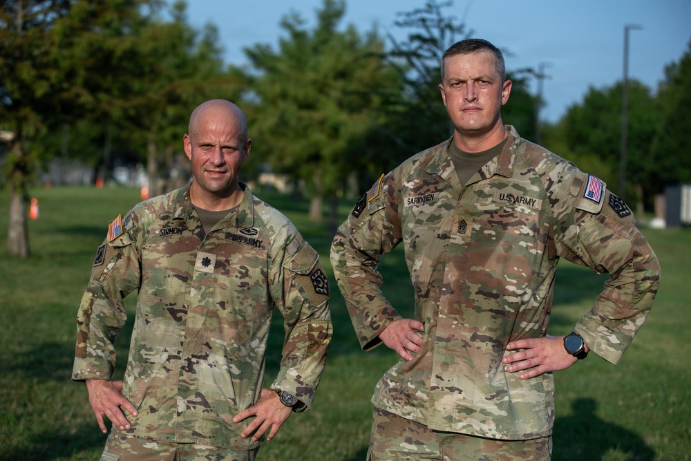 Two U.S. Army soldiers in Operational Camouflage Pattern (OCP) uniforms are posed outdoors on a grassy area, both standing with hands on hips in a confident stance. The soldier on the left has a name tape reading 'SIRMON' and the soldier on the right reads 'SARKKNEN' (or similar), both displaying 'U.S. ARMY' chest tapes and American flag patches. The setting appears to be a military installation during daytime with trees and infrastructure visible in the background.