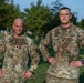 Two U.S. Army soldiers in Operational Camouflage Pattern (OCP) uniforms are posed outdoors on a grassy area, both standing with hands on hips in a confident stance. The soldier on the left has a name tape reading 'SIRMON' and the soldier on the right reads 'SARKKNEN' (or similar), both displaying 'U.S. ARMY' chest tapes and American flag patches. The setting appears to be a military installation during daytime with trees and infrastructure visible in the background.