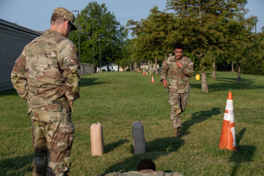 Military personnel in OCP (Operational Camouflage Pattern) uniforms are conducting a physical fitness or obstacle course training exercise on a grassy field. One soldier is actively running through a marked course defined by orange traffic cones and cylindrical obstacles, while another soldier observes from the left. Additional personnel and cones are visible in the background, suggesting a larger organized training event.