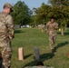 Military personnel in OCP (Operational Camouflage Pattern) uniforms are conducting a physical fitness or obstacle course training exercise on a grassy field. One soldier is actively running through a marked course defined by orange traffic cones and cylindrical obstacles, while another soldier observes from the left. Additional personnel and cones are visible in the background, suggesting a larger organized training event.