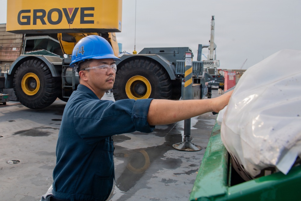 A worker wearing a blue hard hat numbered '14' and safety glasses is handling a plastic-wrapped cargo item on what appears to be a ship's deck or industrial loading area. In the background, a large yellow Grove crane and heavy equipment are visible. The setting suggests a port, shipyard, or heavy industrial cargo operation.