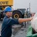 A worker wearing a blue hard hat numbered '14' and safety glasses is handling a plastic-wrapped cargo item on what appears to be a ship's deck or industrial loading area. In the background, a large yellow Grove crane and heavy equipment are visible. The setting suggests a port, shipyard, or heavy industrial cargo operation.