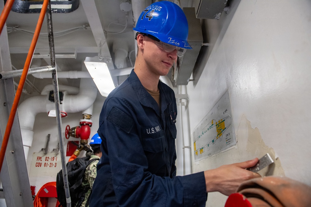 A U.S. Navy sailor wearing a blue hard hat marked '74' and safety glasses is working aboard what appears to be a naval vessel, likely USS John C. Stennis (CVN-74). The sailor is dressed in navy blue coveralls with 'U.S. NAVY' visible on the chest and appears to be involved in maintenance or repair work near a wall-mounted diagram. The interior space shows typical shipboard infrastructure including pipes, valves, and safety equipment.