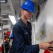 A U.S. Navy sailor wearing a blue hard hat marked '74' and safety glasses is working aboard what appears to be a naval vessel, likely USS John C. Stennis (CVN-74). The sailor is dressed in navy blue coveralls with 'U.S. NAVY' visible on the chest and appears to be involved in maintenance or repair work near a wall-mounted diagram. The interior space shows typical shipboard infrastructure including pipes, valves, and safety equipment.