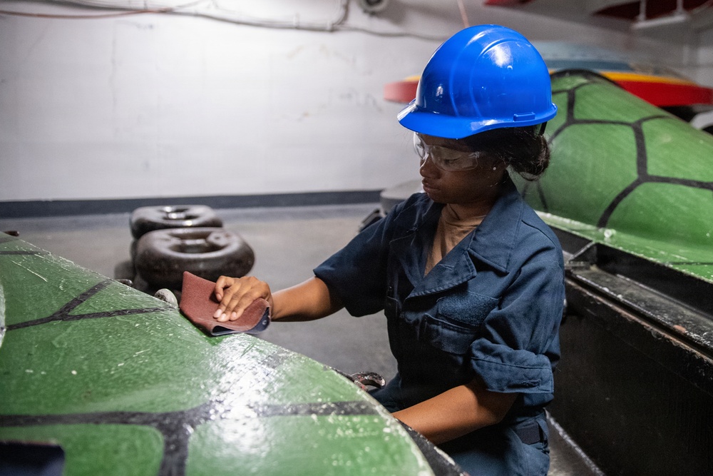 A worker wearing a blue hard hat, safety glasses, and dark navy coveralls is sanding a green painted surface, likely part of a large inflatable boat or rubber vessel. The work is being performed in what appears to be an enclosed industrial or military facility. The task involves surface preparation using sandpaper held by hand.