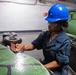 A worker wearing a blue hard hat, safety glasses, and dark navy coveralls is sanding a green painted surface, likely part of a large inflatable boat or rubber vessel. The work is being performed in what appears to be an enclosed industrial or military facility. The task involves surface preparation using sandpaper held by hand.