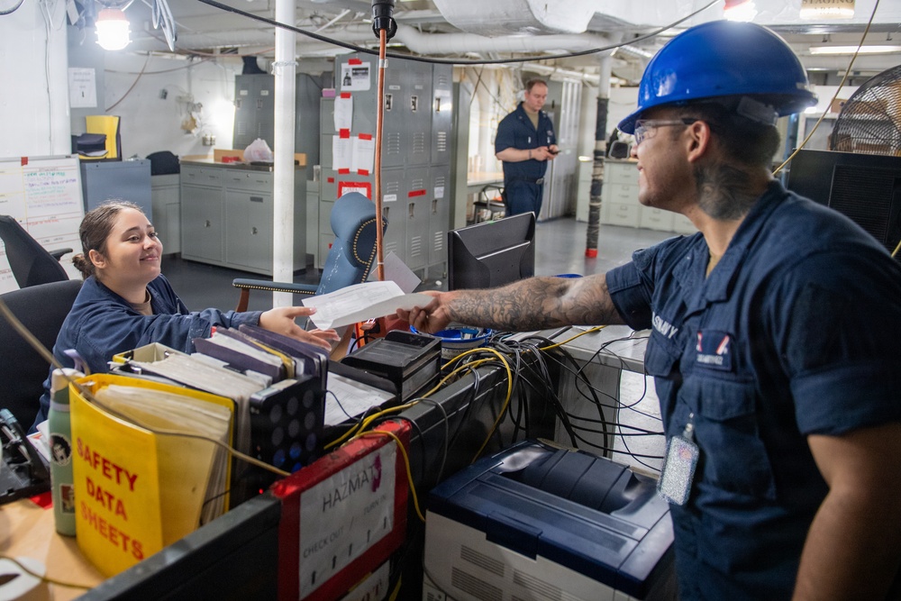 The image shows what appears to be a U.S. Navy shipboard workspace where personnel are exchanging documents across a counter. A female sailor seated at a desk is smiling while handing or receiving paperwork from a male sailor wearing a blue hard hat and safety glasses. A third sailor is visible in the background near gray storage lockers.