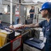 The image shows what appears to be a U.S. Navy shipboard workspace where personnel are exchanging documents across a counter. A female sailor seated at a desk is smiling while handing or receiving paperwork from a male sailor wearing a blue hard hat and safety glasses. A third sailor is visible in the background near gray storage lockers.