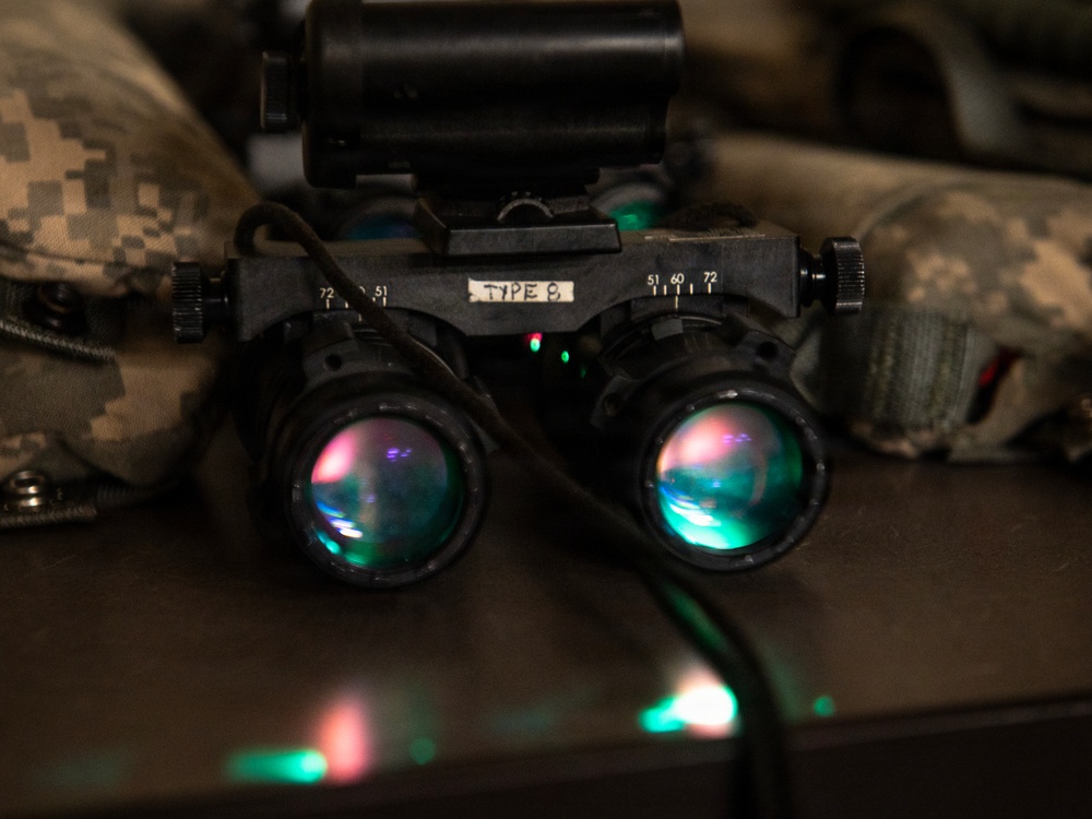 The image shows a pair of military night vision goggles (NVGs) labeled 'TYPE 8' resting on a dark surface. The dual-tube optical device features multi-coated lenses with visible teal and pink light reflections, along with small red and green indicator LEDs. Military camouflage gear (ACU/UCP pattern) is visible in the background.