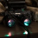 The image shows a pair of military night vision goggles (NVGs) labeled 'TYPE 8' resting on a dark surface. The dual-tube optical device features multi-coated lenses with visible teal and pink light reflections, along with small red and green indicator LEDs. Military camouflage gear (ACU/UCP pattern) is visible in the background.