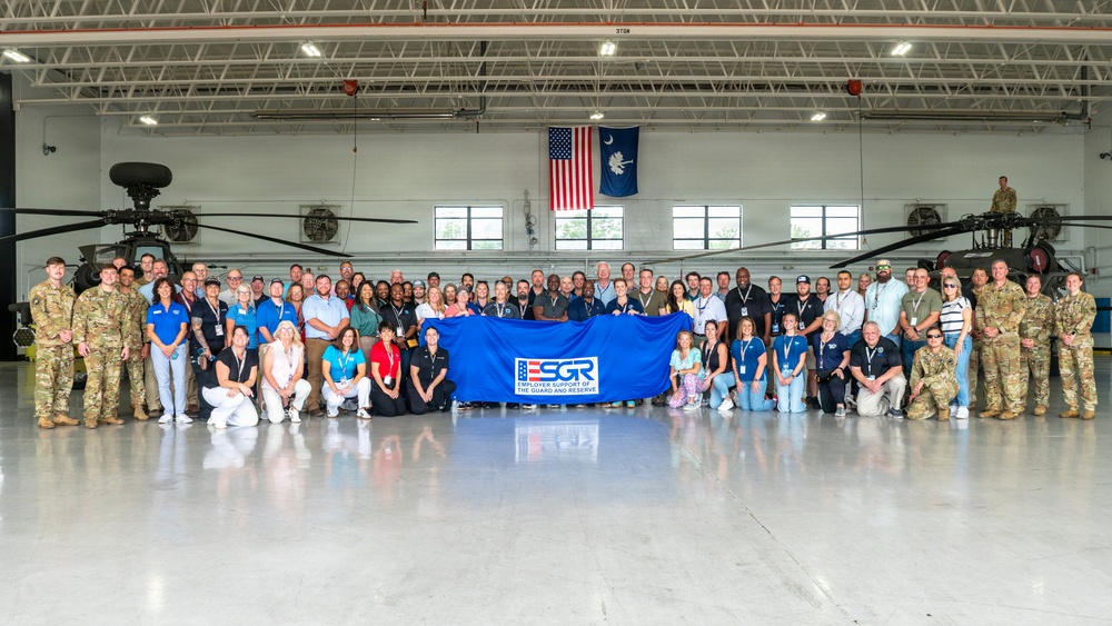 A large group of military personnel and civilians pose for a formal group photo inside a military helicopter hangar. They are holding a blue ESGR (Employer Support of the Guard and Reserve) banner, indicating this is an official ESGR event. Two military helicopters (appearing to be Apache attack helicopters) are visible in the background, along with an American flag and a South Carolina state flag.