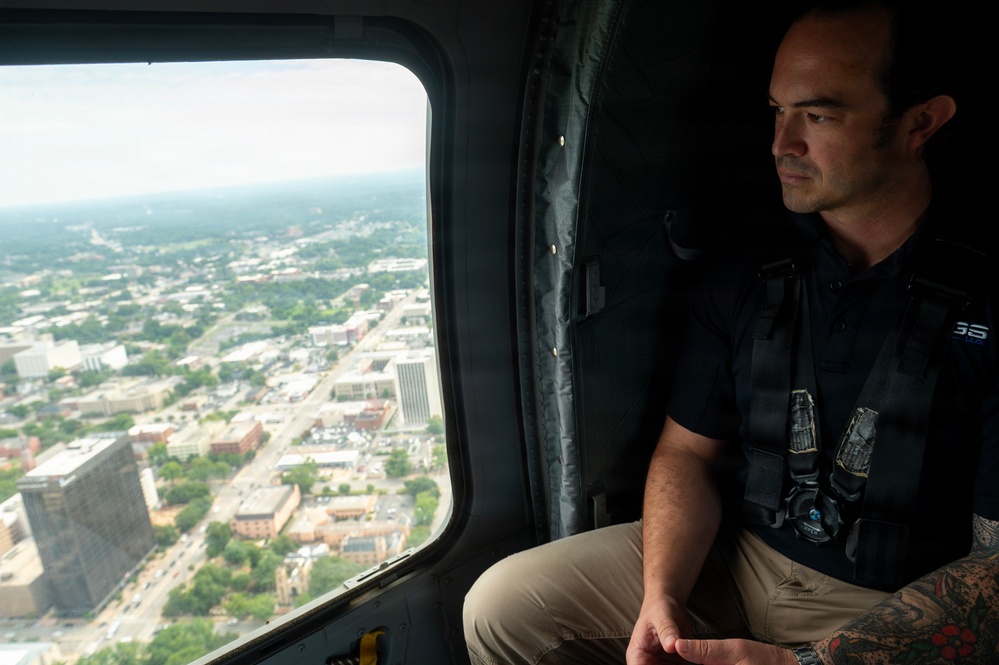 A man wearing a tactical vest and dark polo shirt sits inside a helicopter, looking out a large window at an aerial view of an urban cityscape below. The city visible through the window shows a mix of commercial buildings, streets, and tree-lined areas, suggesting a mid-sized American city. The man appears to be on some form of official or operational flight, indicated by his tactical gear and harness equipment.