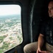 A man wearing a tactical vest and dark polo shirt sits inside a helicopter, looking out a large window at an aerial view of an urban cityscape below. The city visible through the window shows a mix of commercial buildings, streets, and tree-lined areas, suggesting a mid-sized American city. The man appears to be on some form of official or operational flight, indicated by his tactical gear and harness equipment.