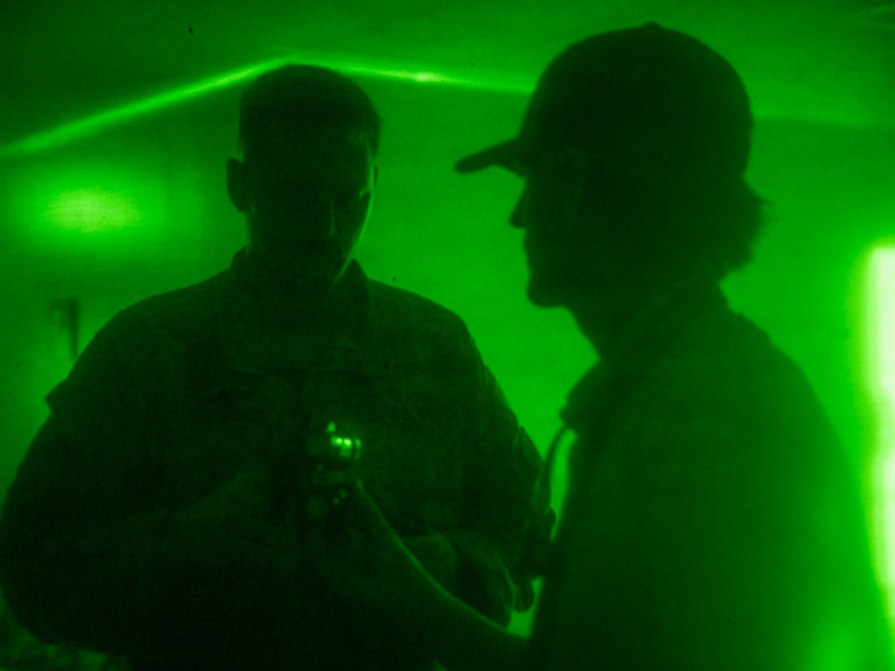 Two individuals are silhouetted against an intense green light environment, likely illuminated by laser or specialized lighting. One person on the left appears to be holding a small device emitting green light, while the person on the right wears a baseball cap. The scene has a low-visibility, atmospheric quality consistent with a training exercise, nightclub, or tactical environment.