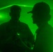 Two individuals are silhouetted against an intense green light environment, likely illuminated by laser or specialized lighting. One person on the left appears to be holding a small device emitting green light, while the person on the right wears a baseball cap. The scene has a low-visibility, atmospheric quality consistent with a training exercise, nightclub, or tactical environment.