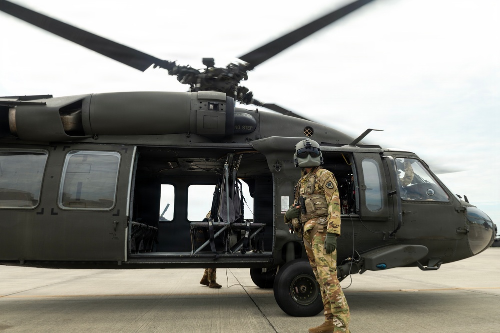 A military helicopter, identifiable as a UH-60 Black Hawk, is shown on a tarmac or flight line with its rotor blades in motion. A soldier in full combat gear including a flight helmet and OCP (Operational Camouflage Pattern) uniform stands beside the open side door of the aircraft. A second personnel member is partially visible near the rear of the helicopter.