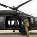 A military helicopter, identifiable as a UH-60 Black Hawk, is shown on a tarmac or flight line with its rotor blades in motion. A soldier in full combat gear including a flight helmet and OCP (Operational Camouflage Pattern) uniform stands beside the open side door of the aircraft. A second personnel member is partially visible near the rear of the helicopter.