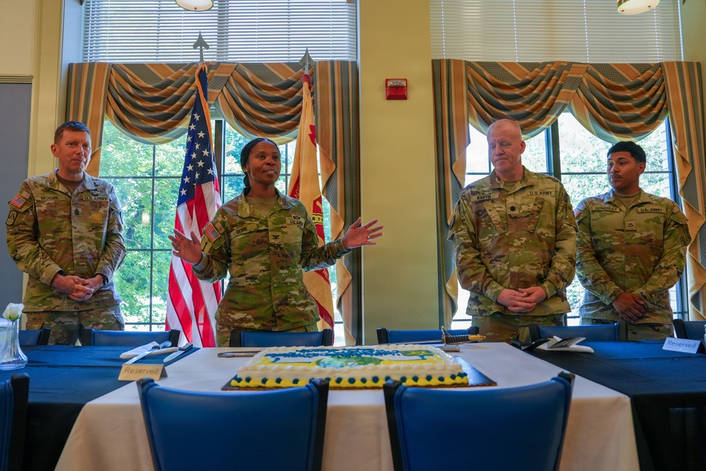 Four U.S. Army soldiers in OCP (Operational Camouflage Pattern) uniforms stand behind a table at what appears to be a formal military ceremony or celebration. A decorated sheet cake is visible on the table in the foreground, and an American flag along with what appears to be a unit flag are displayed in the background. One female soldier appears to be speaking or addressing the group.