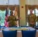Four U.S. Army soldiers in OCP (Operational Camouflage Pattern) uniforms stand behind a table at what appears to be a formal military ceremony or celebration. A decorated sheet cake is visible on the table in the foreground, and an American flag along with what appears to be a unit flag are displayed in the background. One female soldier appears to be speaking or addressing the group.