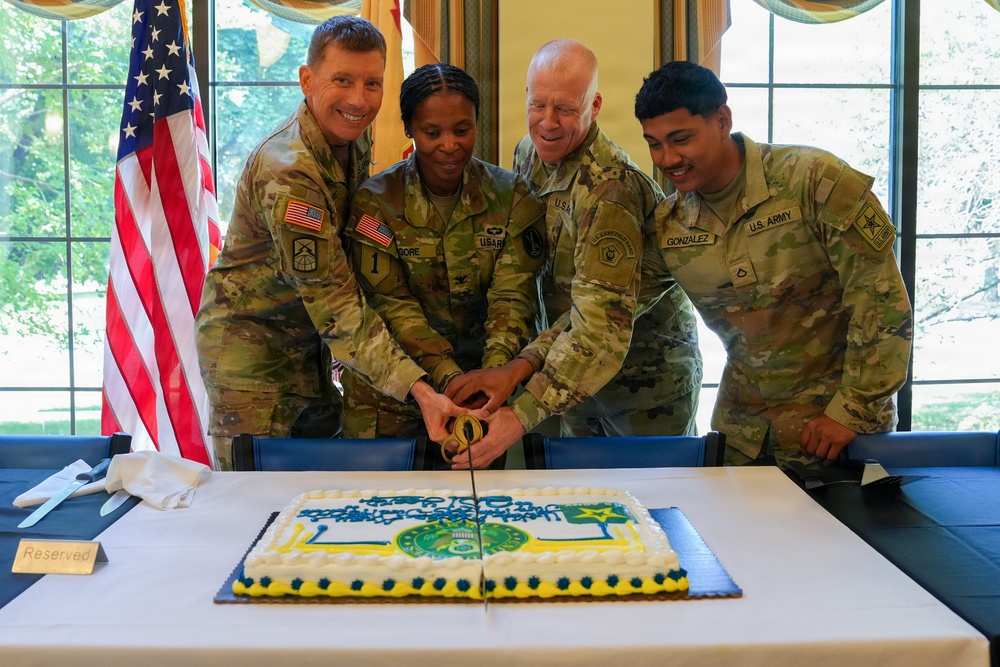 Four U.S. Army soldiers in OCP (Operational Camouflage Pattern) uniforms are jointly cutting a decorated military cake at what appears to be a formal ceremony. The cake features blue, yellow, and green decorations with what appears to be a unit insignia. An American flag and what appears to be a unit guidon are visible in the background.