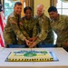 Four U.S. Army soldiers in OCP (Operational Camouflage Pattern) uniforms are jointly cutting a decorated military cake at what appears to be a formal ceremony. The cake features blue, yellow, and green decorations with what appears to be a unit insignia. An American flag and what appears to be a unit guidon are visible in the background.