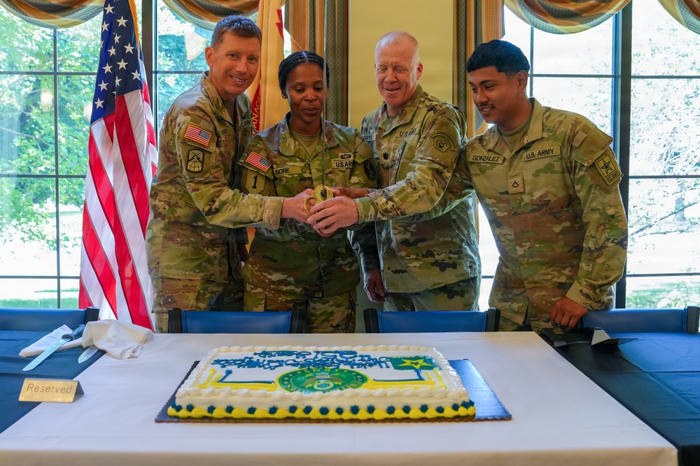 Four U.S. Army soldiers in OCP (Operational Camouflage Pattern) uniforms are gathered around a decorated military cake, performing a ceremonial cake-cutting. The cake features what appears to be a U.S. Army branch or unit insignia in blue, green, and yellow. An American flag and what appears to be a unit guidon are visible in the background.