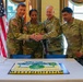 Four U.S. Army soldiers in OCP (Operational Camouflage Pattern) uniforms are gathered around a decorated military cake, performing a ceremonial cake-cutting. The cake features what appears to be a U.S. Army branch or unit insignia in blue, green, and yellow. An American flag and what appears to be a unit guidon are visible in the background.