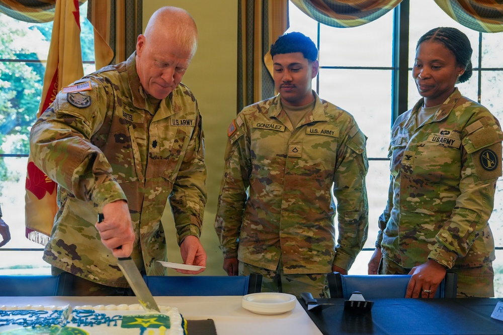 Three U.S. Army soldiers in OCP (Operational Camouflage Pattern) uniforms are participating in a cake-cutting ceremony, likely a military birthday or unit celebration. The senior soldier on the left, identified by his name tape as 'ARTEL,' is cutting a decorated sheet cake with a large ceremonial sword/saber. Two other soldiers, GONZALEZ and a female soldier, observe the cutting.