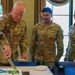 Three U.S. Army soldiers in OCP (Operational Camouflage Pattern) uniforms are participating in a cake-cutting ceremony, likely a military birthday or unit celebration. The senior soldier on the left, identified by his name tape as 'ARTEL,' is cutting a decorated sheet cake with a large ceremonial sword/saber. Two other soldiers, GONZALEZ and a female soldier, observe the cutting.