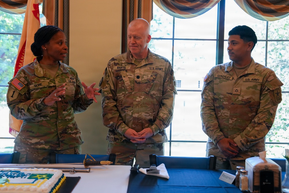 Three U.S. Army soldiers in OCP (Operational Camouflage Pattern) uniforms are gathered around a table at what appears to be a military ceremony or celebration. A decorated cake and a ceremonial sword are visible on the table in the foreground, suggesting this may be a unit anniversary or promotion event. The soldier on the left, identified as 'Gore,' appears to be speaking while the other two, 'Bartel' and 'Gonzalez,' listen attentively.