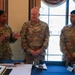 Three U.S. Army soldiers in OCP (Operational Camouflage Pattern) uniforms are gathered around a table at what appears to be a military ceremony or celebration. A decorated cake and a ceremonial sword are visible on the table in the foreground, suggesting this may be a unit anniversary or promotion event. The soldier on the left, identified as 'Gore,' appears to be speaking while the other two, 'Bartel' and 'Gonzalez,' listen attentively.