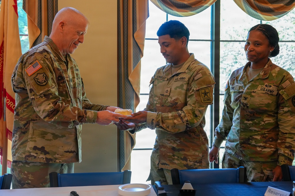 Three U.S. Army soldiers in OCP (Operational Camouflage Pattern) uniforms are gathered in what appears to be a formal dining or meeting room. A senior soldier on the left is receiving or exchanging a small food item from a soldier identified by nametape as 'GONZALEZ,' while a third soldier identified as 'MOORE' observes with a smile. The setting suggests a ceremonial or recognition event.