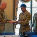 Three U.S. Army soldiers in OCP (Operational Camouflage Pattern) uniforms are gathered in what appears to be a formal dining or meeting room. A senior soldier on the left is receiving or exchanging a small food item from a soldier identified by nametape as 'GONZALEZ,' while a third soldier identified as 'MOORE' observes with a smile. The setting suggests a ceremonial or recognition event.