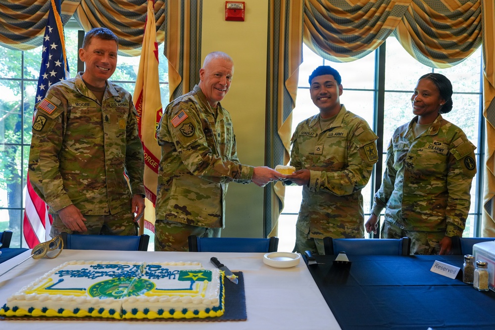 Four U.S. Army soldiers in OCP (Operational Camouflage Pattern) uniforms are gathered around a table during what appears to be a military celebration or ceremony. A senior officer is handing a piece of cake to a junior enlisted soldier identified as 'Gonzalez,' while soldier 'Moore' stands to the left and a female soldier stands to the right. A decorated sheet cake with a military insignia is visible on the table in the foreground.