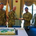 Four U.S. Army soldiers in OCP (Operational Camouflage Pattern) uniforms are gathered around a table during what appears to be a military celebration or ceremony. A senior officer is handing a piece of cake to a junior enlisted soldier identified as 'Gonzalez,' while soldier 'Moore' stands to the left and a female soldier stands to the right. A decorated sheet cake with a military insignia is visible on the table in the foreground.